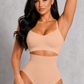 A woman modeling a beige two-piece shapewear set consisting of a bralette and high-waisted briefs with a midriff cutout, set against a textured gray studio background.