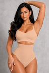 A woman modeling a beige two-piece shapewear set consisting of a bralette and high-waisted briefs with a midriff cutout, set against a textured gray studio background.