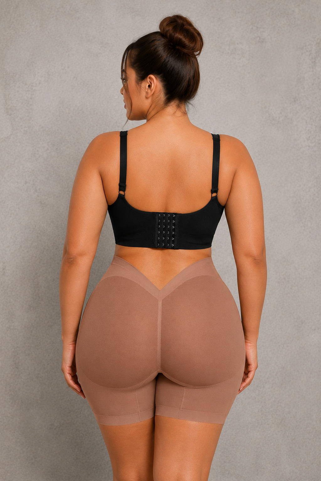 A woman shown from the back wearing a black bra and high-waisted shapewear shorts in a nude beige color, standing against a textured gray wall.