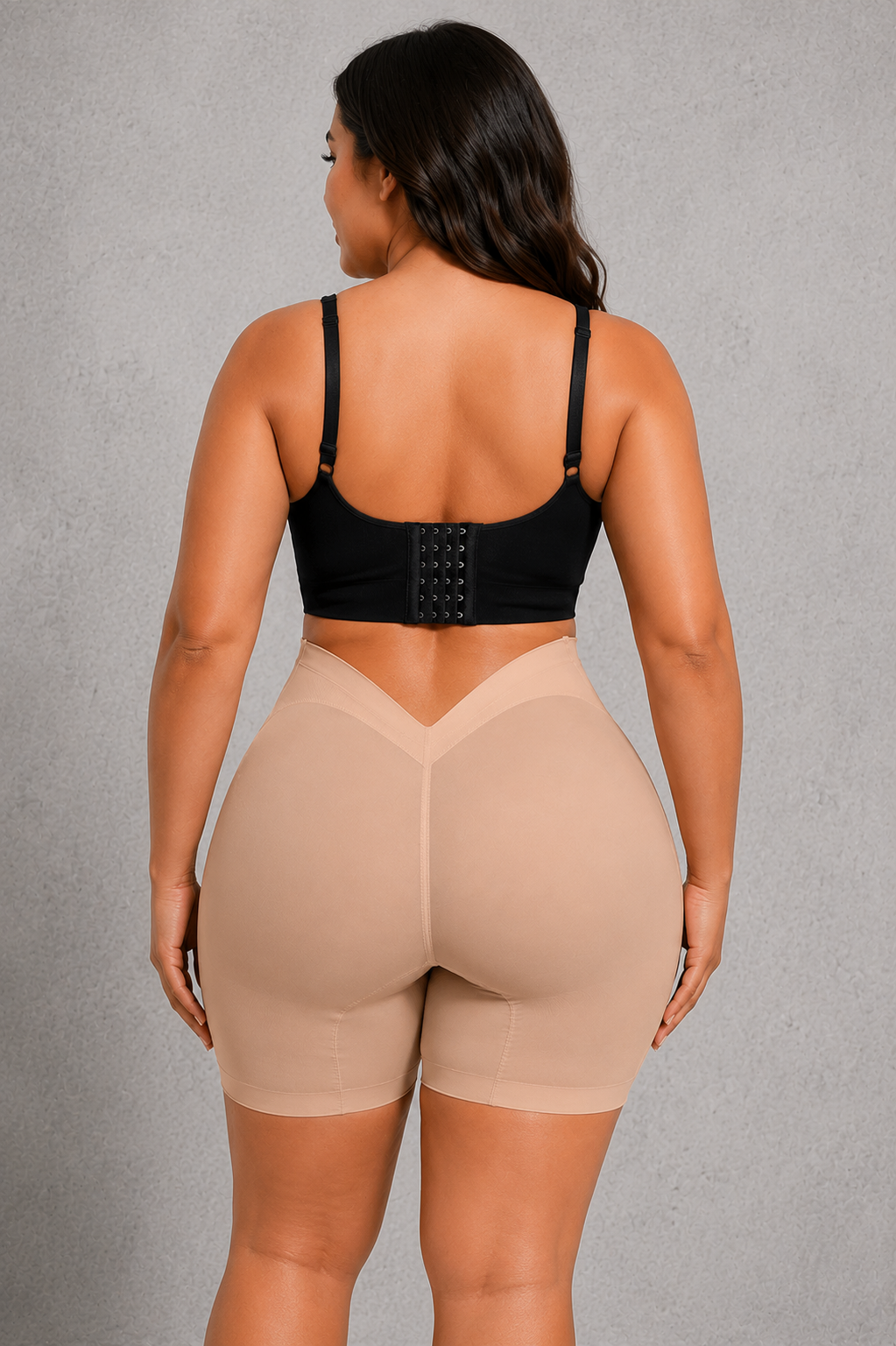 A woman shown from the back wearing beige high-waisted shapewear shorts and a black strappy bra, standing against a textured gray studio background.