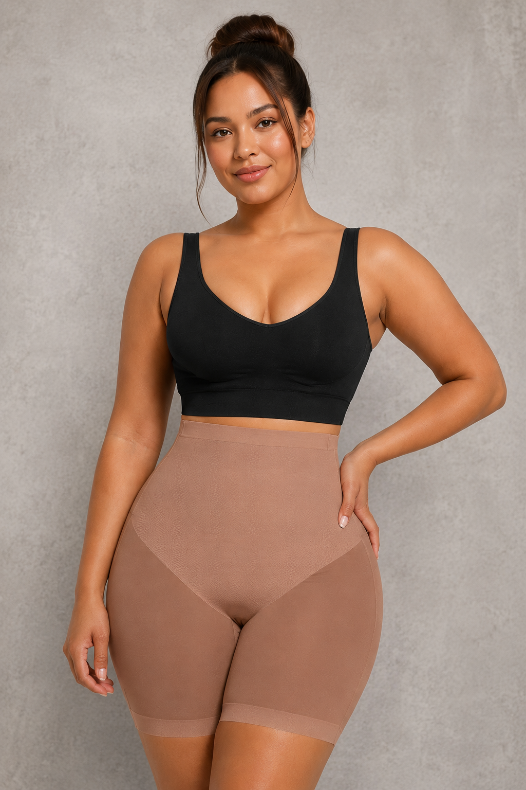 A plus-size woman modeling a black scoop-neck crop top and high-waisted shapewear shorts in a nude shade, standing against a textured gray studio background.