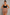 A woman modeling a high-waisted, skin-toned shapewear brief with a black sports bra, holding the waistband outwards to demonstrate its stretch and fit, against a textured gray studio background.