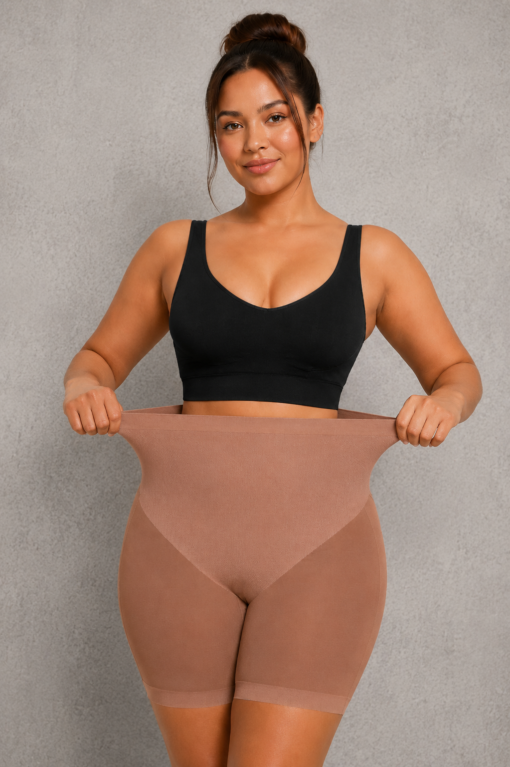 A woman modeling a high-waisted, skin-toned shapewear brief with a black sports bra, holding the waistband outwards to demonstrate its stretch and fit, against a textured gray studio background.