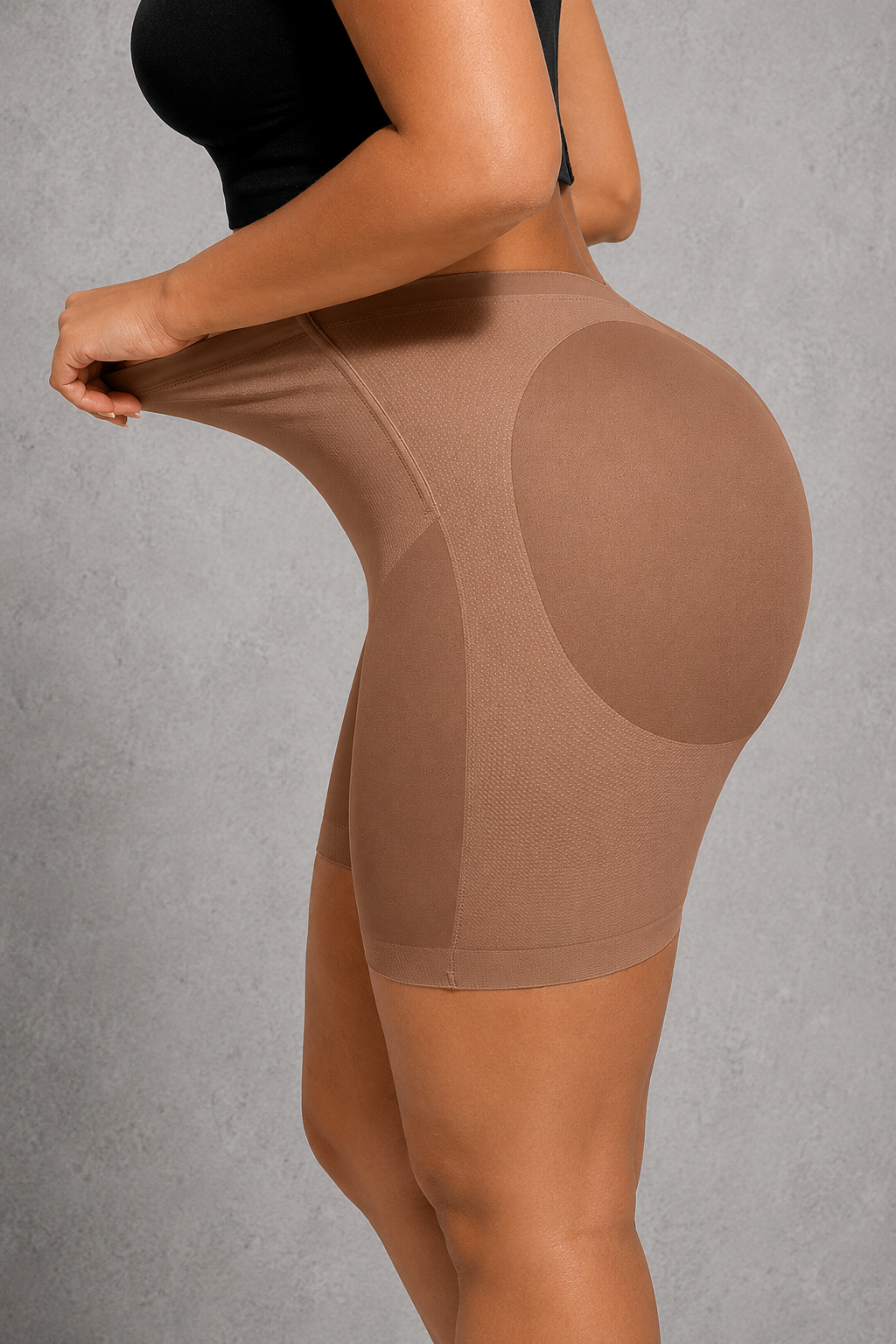 A model wearing a beige high-waisted compression shapewear brief, lifting the left side of the waist to demonstrate support, against a textured gray background.