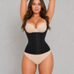 A woman modeling a black structured waist trainer corset over a beige bra and matching beige underwear, standing against a plain light gray studio background with arms raised overhead.