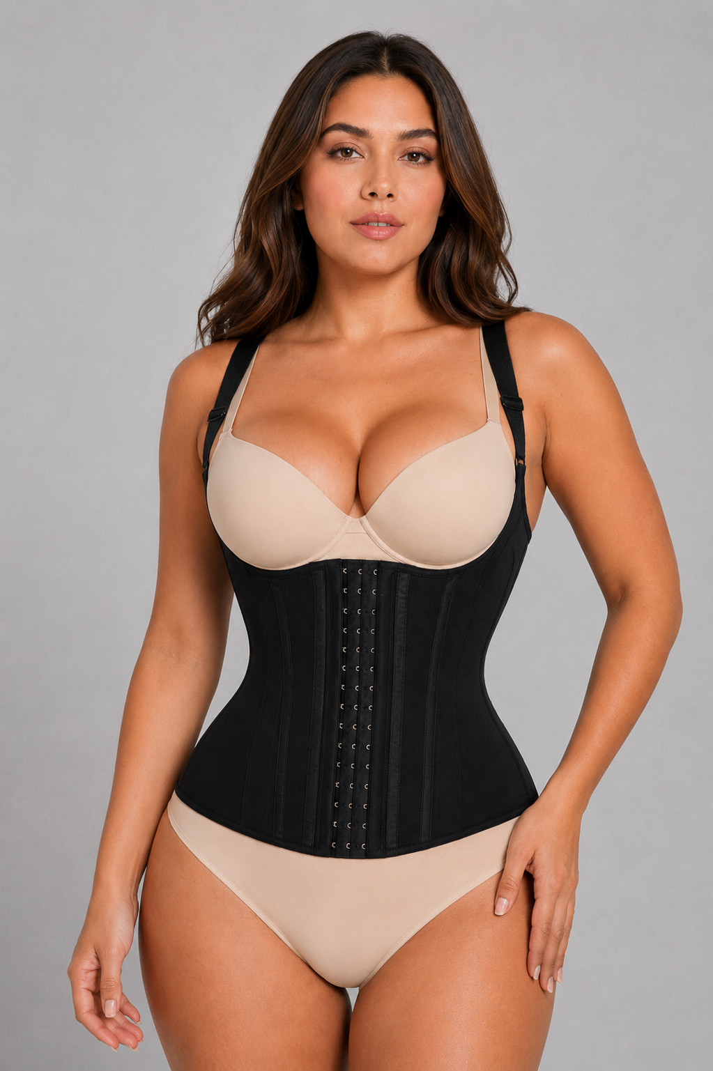 A woman modeling a black structured waist trainer corset over a beige bra and matching beige underwear, photographed against a plain light gray studio background.