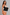 A woman modeling a black waist trainer corset over beige underwear, holding a matching black panel against a plain light gray studio background.