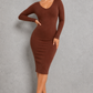 A woman modeling a long-sleeve, bodycon midi dress in a rich brown color, standing against a textured gray studio wall.