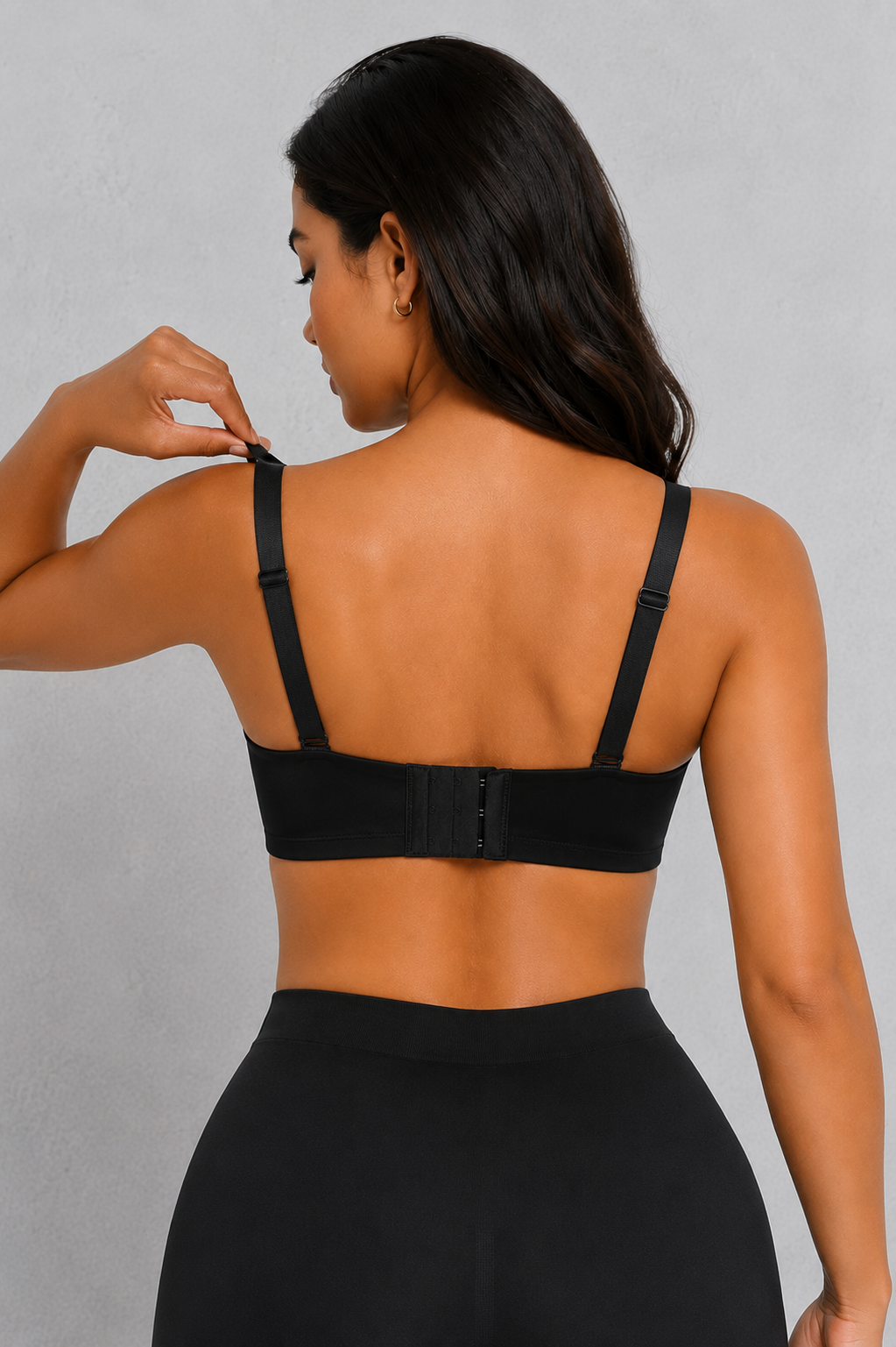 A woman shown from the back wearing a black bralette with adjustable shoulder straps and a hook-and-eye back closure, paired with black bottoms, against a light gray textured background.