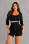 A woman modeling a black two-piece shapewear set consisting of a cropped long-sleeve top and matching high-waisted shorts with lace trim and front lace-up detailing, set against a textured gray studio background.
