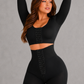 A woman modeling a black two-piece activewear set consisting of a long-sleeve crop top and high-waisted shorts with lace trim and lace-up front details, set against a plain gray studio background.