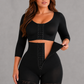 A woman modeling a black two-piece shapewear set consisting of a long-sleeve crop top and matching high-waisted shorts with lace trim and front hook-and-eye closure, against a solid gray studio background.