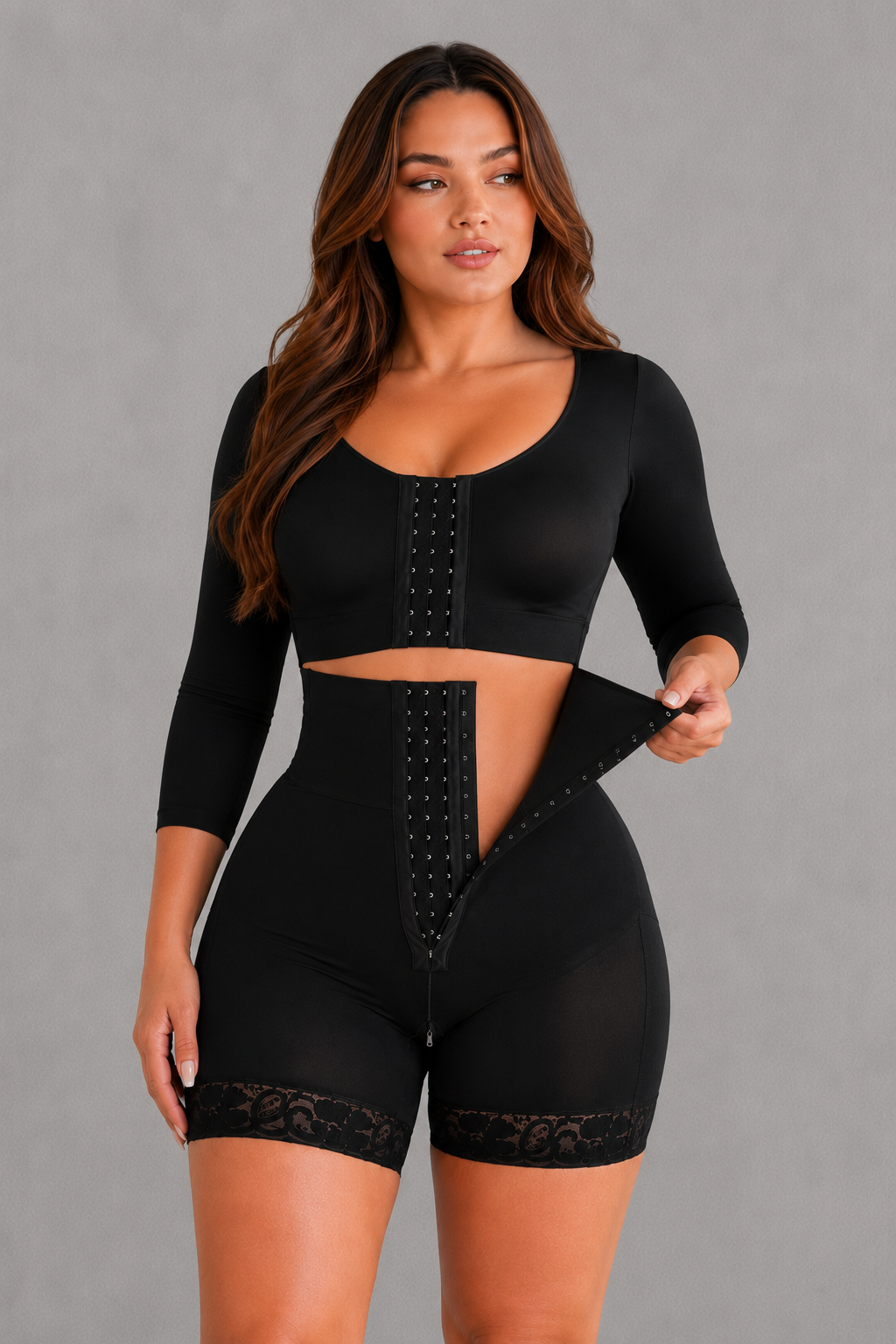 A woman modeling a black two-piece shapewear set consisting of a long-sleeve crop top and matching high-waisted shorts with lace trim and front hook-and-eye closure, against a solid gray studio background.