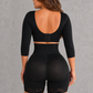 A woman shown from the back wearing a black three-piece shapewear set consisting of a long-sleeve crop top with lace-up back detail, a high-waisted waist trainer, and matching shapewear shorts with lace trim, set against a textured gray background.