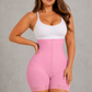 A woman modeling a pink high-waisted shapewear brief with lace trim and a front button closure, paired with a white tank top, against a textured gray studio background.