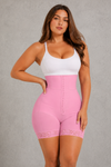 A woman modeling a pink high-waisted shapewear brief with lace trim and a front button closure, paired with a white tank top, against a textured gray studio background.