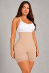 A woman modeling a beige high-waisted shapewear bodysuit with a white camisole top, standing against a textured gray studio background.