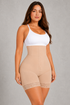 A woman modeling a beige high-waisted shapewear bodysuit with a white camisole top, standing against a textured gray studio background.
