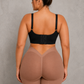 A woman shown from the back wearing a black bra and high-waisted shapewear shorts in a nude beige color, standing against a textured gray wall.