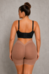 A woman shown from the back wearing a black bra and high-waisted shapewear shorts in a nude beige color, standing against a textured gray wall.
