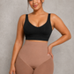 A plus-size woman modeling a black scoop-neck crop top and high-waisted shapewear shorts in a nude shade, standing against a textured gray studio background.