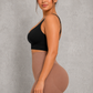 A woman posing in a side profile view, wearing a black sports bra and high-waisted, skin-toned shapewear shorts against a textured gray studio background.