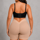 A woman shown from the back wearing beige high-waisted shapewear shorts and a black strappy bra, standing against a textured gray studio background.