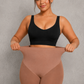 A woman modeling a high-waisted, skin-toned shapewear brief with a black sports bra, holding the waistband outwards to demonstrate its stretch and fit, against a textured gray studio background.