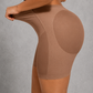 A model wearing a beige high-waisted compression shapewear brief, lifting the left side of the waist to demonstrate support, against a textured gray background.