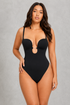 A woman modeling a black one-piece bodysuit with a keyhole cutout at the bust, thin shoulder straps, and high-cut legs, posed against a textured gray studio background.
