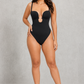 A woman modeling a black one-piece swimsuit with a keyhole cutout at the center, set against a textured gray studio backdrop.