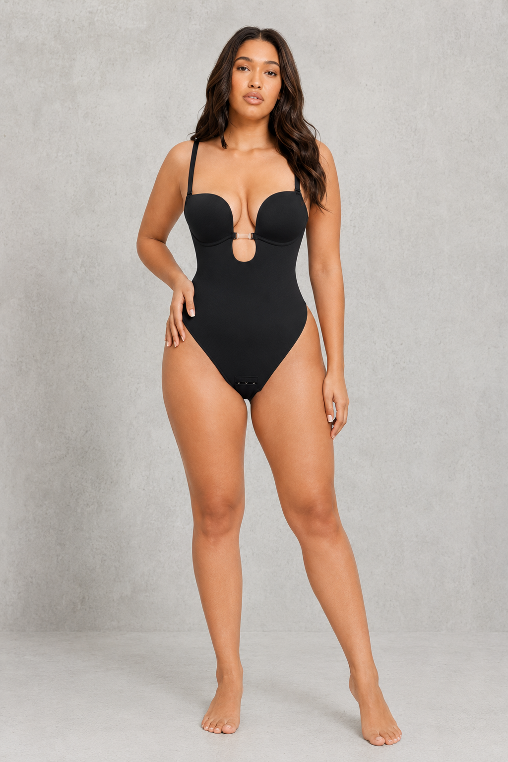 A woman modeling a black one-piece swimsuit with a keyhole cutout at the center, set against a textured gray studio backdrop.